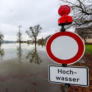 Starkregen, Sturzfluten, Hochwasser, Dürre: Die Gefahren durch Extremwetter steigen. Wie gut ist Baden-Württemberg vorbereitet? Wie steht es um den Hochwasserschutz, wie um Renaturierung und Katastrophenschutz? Darüber sprechen der Katastrophenforscher Andreas Schäfer und der Oberbürgermeister von Gaggenau, Michael Pfeiffer, in SWR1 Leute. Das Symbolbild zeigt ein Schild "Durchfahrt verboten"und ein Hochwasser-Hinweisschild, dahinter stehen Bäume im Wasser.a