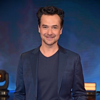 Michael Kaeshammer steht im blauen Anzug vor einem blauen Hintergrund und lacht in die Kamera