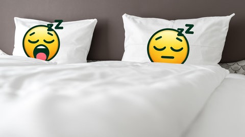Ein Bett mit zwei weißen Decken und zwei weißen Kissen mit zwei schlafenden Emojigesichern. (Foto: picture-alliance / Reportdienste, CHROMORANGE | Michael Bihlmayer) Ein Bett mit zwei weißen Decken und zwei weißen Kissen mit zwei schlafenden Emojigesichern.