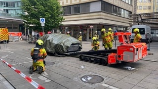 Feuerwehrleute transportieren ein abgedecktes E-Auto nach einem Brand in einer Tiefgarage mit einem speziellen Gerät ab.