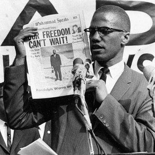 Malcolm X hält bei einer Demonstration 1963 eine Zeitung in die Höhe. Als Schlagzeile steht darauf: „Our freedom can’t wait.“ – „Unsere Freiheit kann nicht warten.“