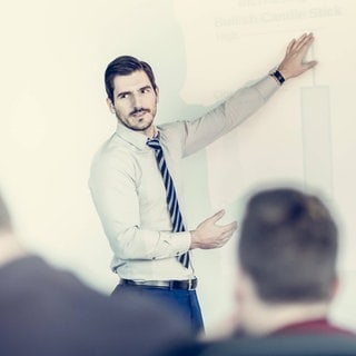 Ein Mann erklärt während einer Präsentation: Der Begriff "Powerpoint-Genau" stammt von dem Freiburger Linguisten Peter Auer. Es ist ein junges sprachliches Phänomen, das häufig in Vorträgen oder Prüfungssituationen auftaucht. 